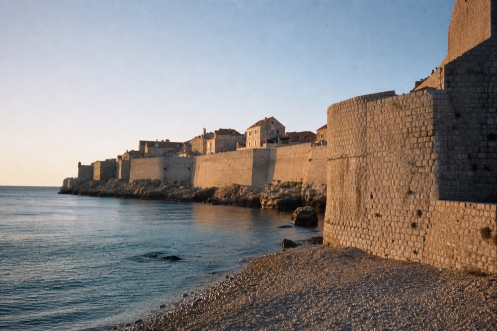 A stone city wall rising above a pebble shoreline and calm blue sea
