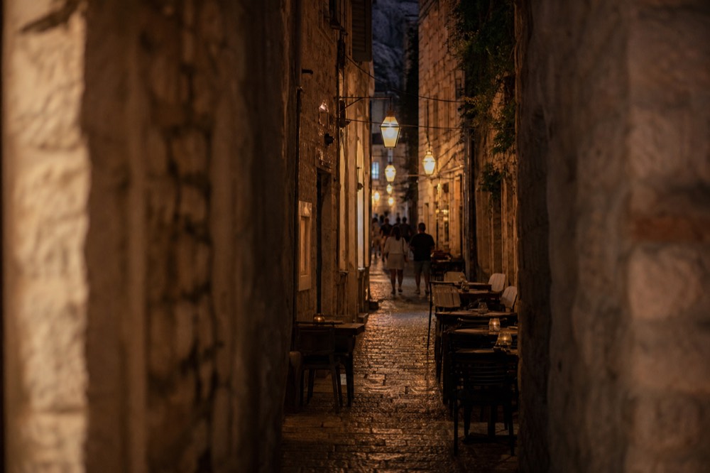 A lantern lit stone alley in Dubrovnik Old Town at night with cafe tables set out along the lane