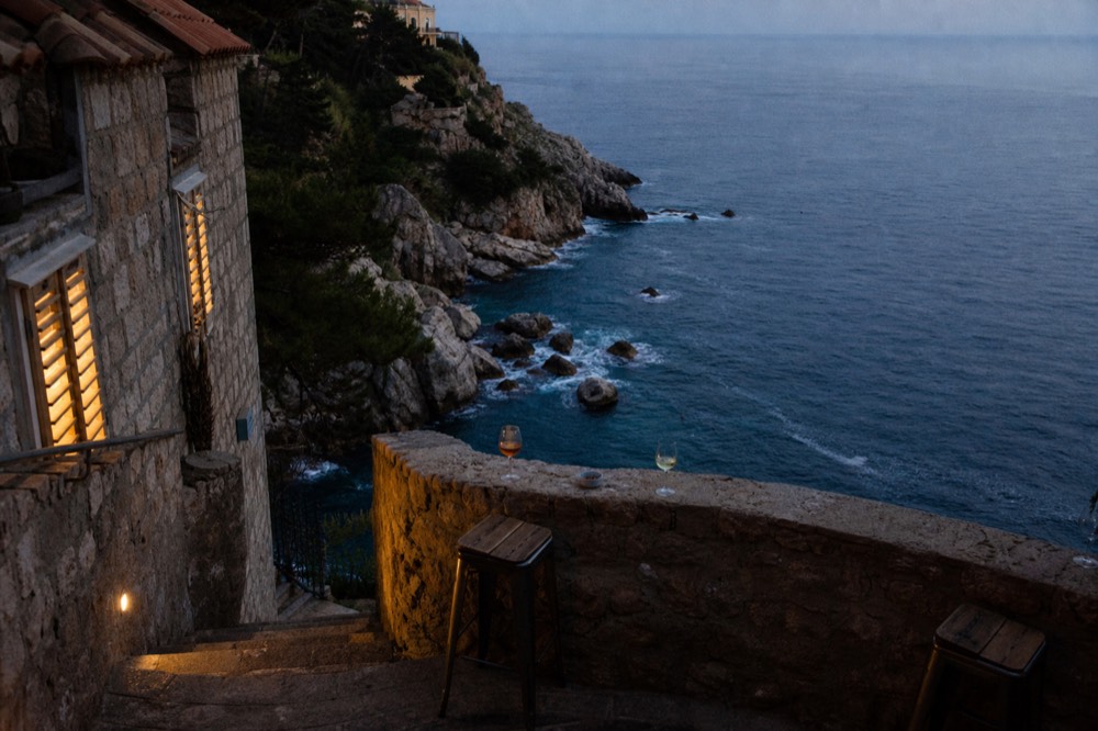 A simple cliffside bar terrace with drinks on a low wall and the sea and rocks below