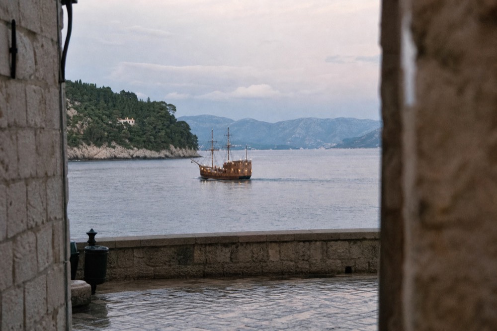 A wooden sailing ship with masts and lanterns cruising on calm evening water near a coastline