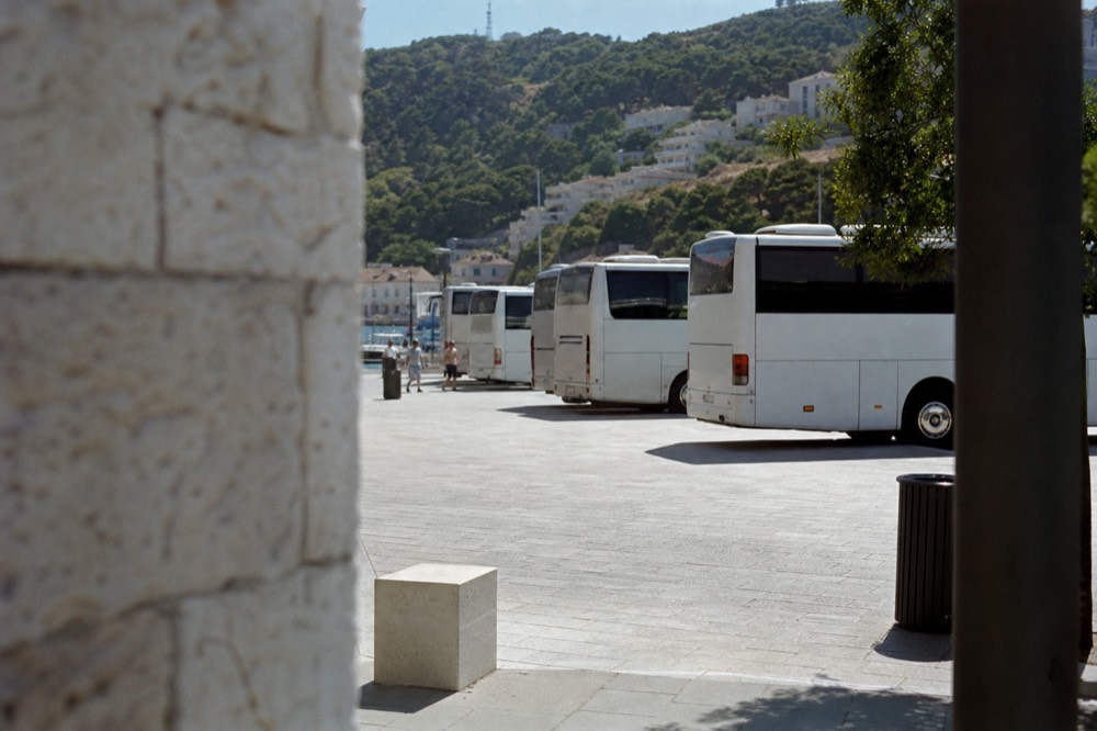 A row of regional buses at a coastal station with hills rising behind the port