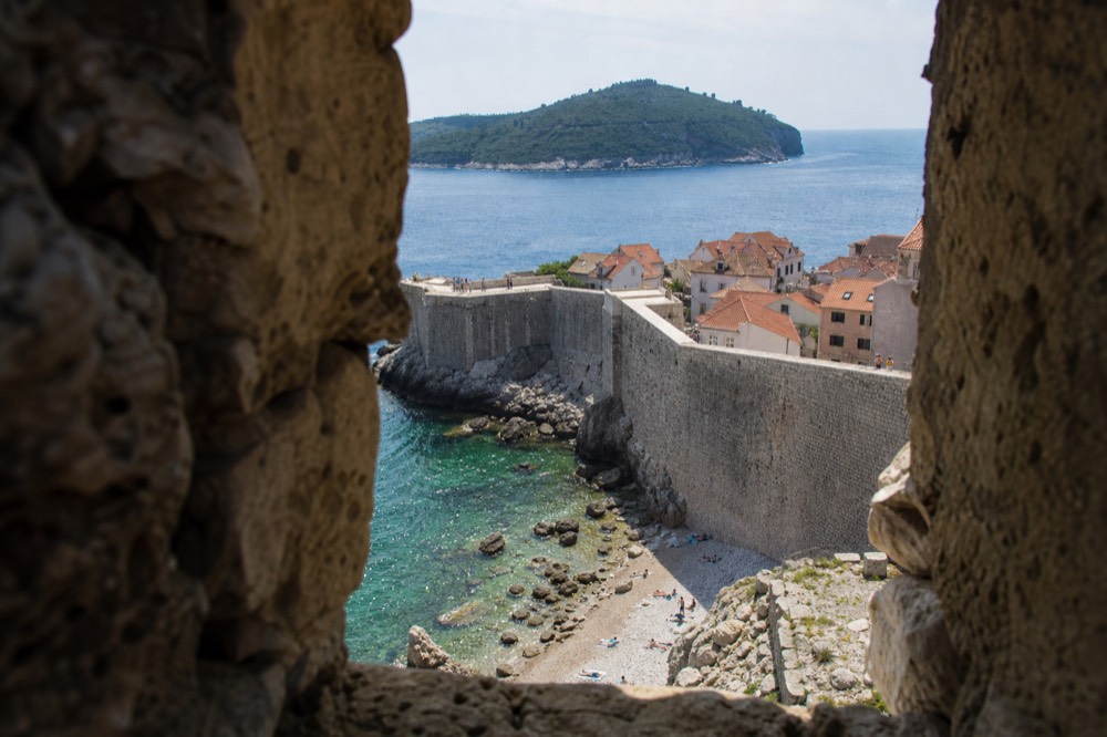 Stone fortification walls curving above red rooftops with the sea and an island beyond