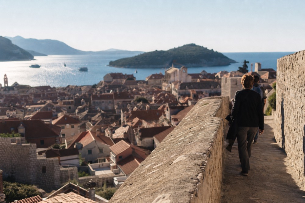 Walkers on a stone wall path above tightly packed red roofs under a clear sky