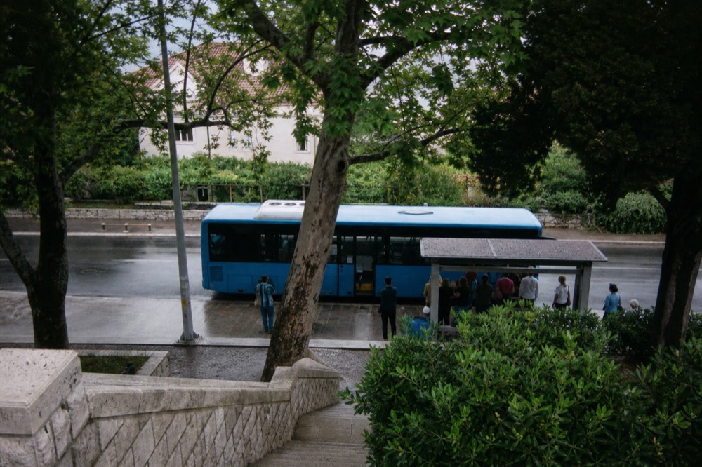 A blue local bus at a stop on a tree-lined street in Lapad with passengers waiting in the shade