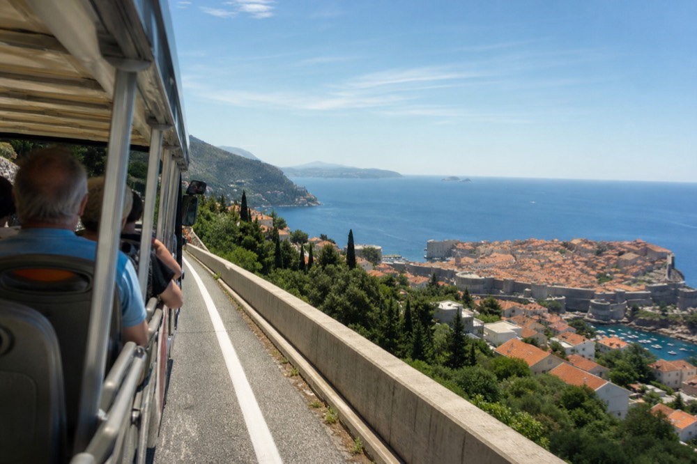 An open-sided panorama bus driving along a coastal road with the Adriatic sea and Dubrovnik below