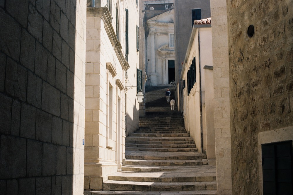 A stone staircase inside an old town leading up between pale buildings toward a church facade