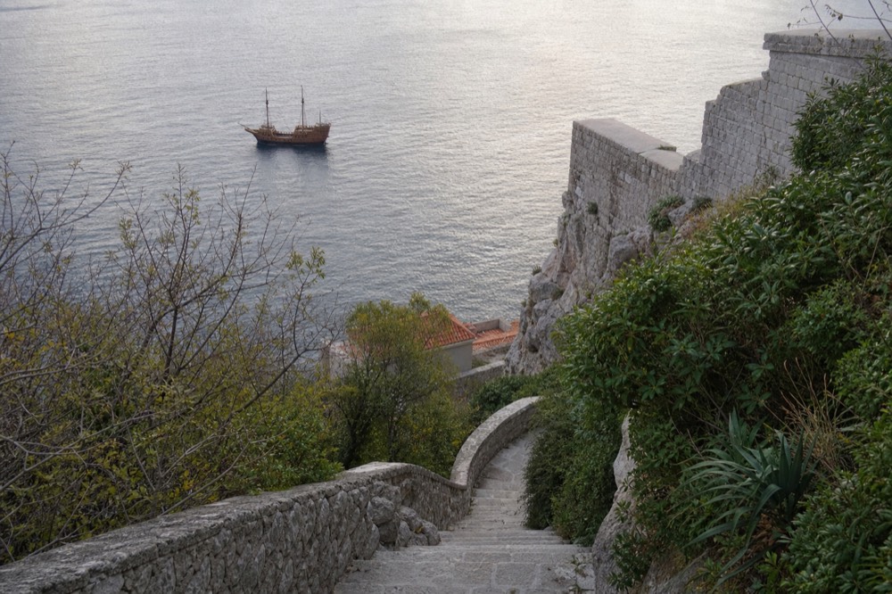 A wooden sailing ship silhouetted on calm evening water below stone walls