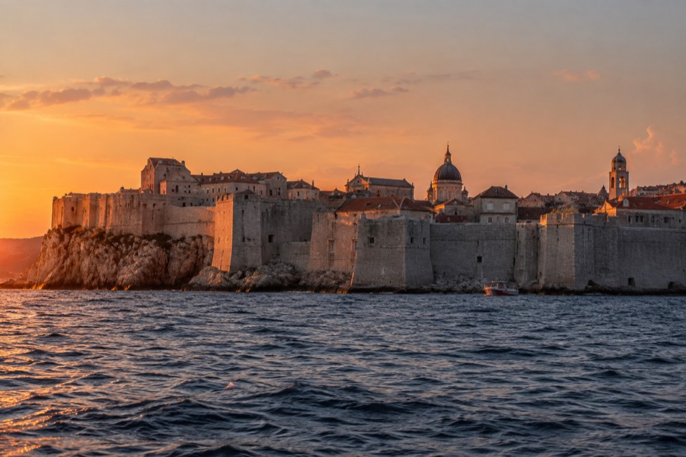 A wide view of a walled old town from the sea at dusk with warm light on stone towers