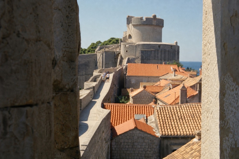 Medieval stone wall walkway curving above terracotta roofs toward a round tower