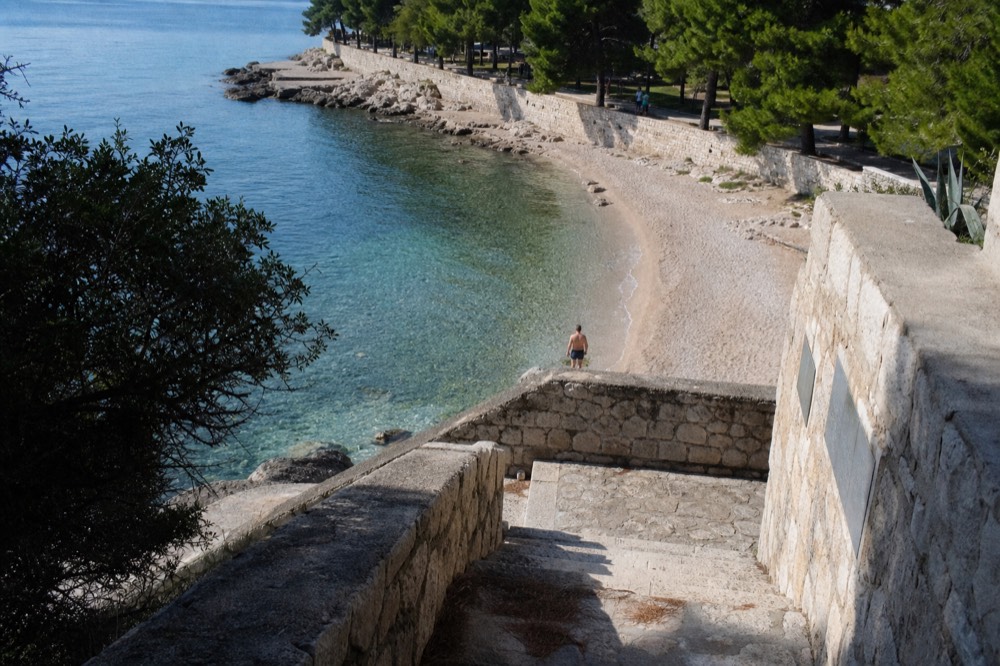 A curved sandy beach with calm shallow water and pine trees along a seaside promenade
