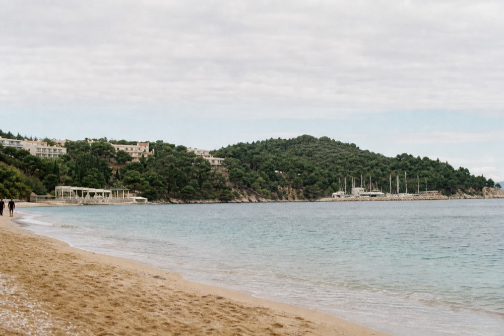 A view along a sandy beach toward pine-covered headlands and a small marina