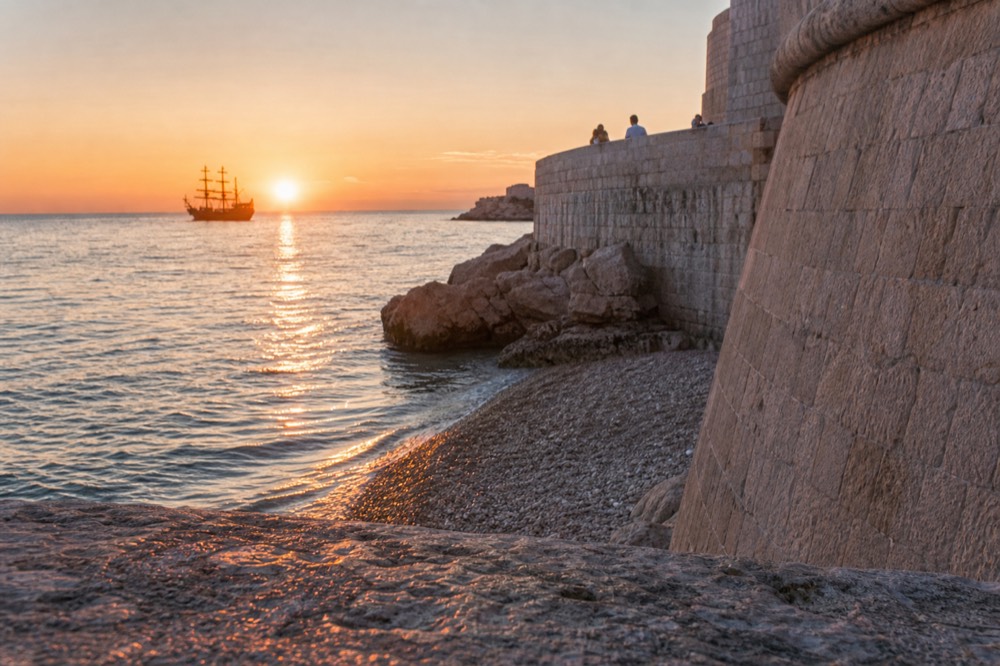 A wooden tall ship silhouetted against an orange sunset over the sea