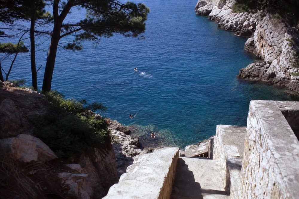 Swimmers in clear blue water below rocky cliffs on a Dubrovnik island shoreline