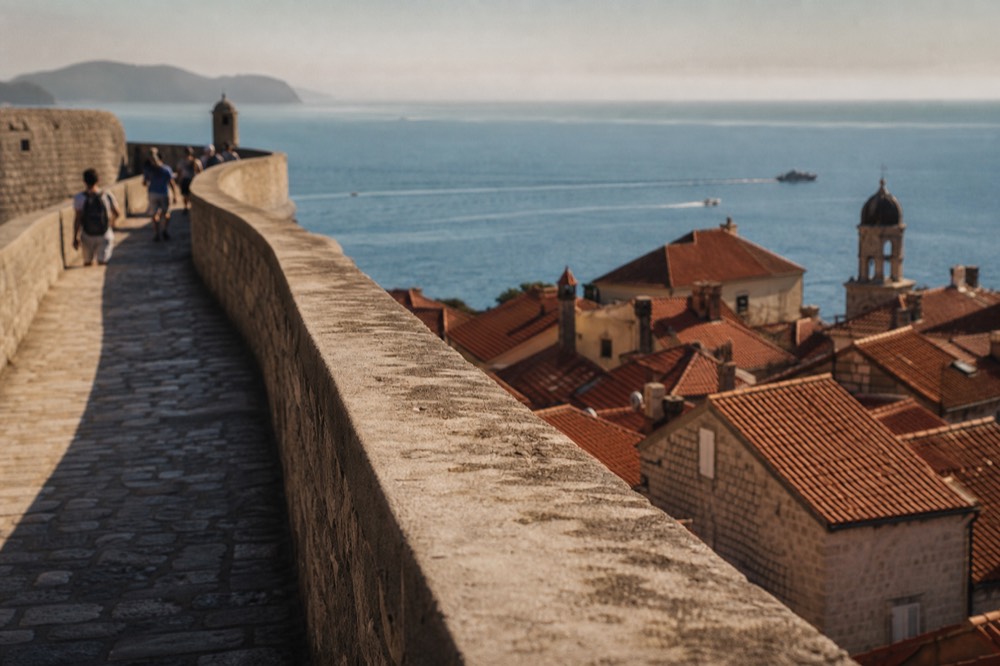 City walls walkway curving above red rooftops toward the blue Adriatic