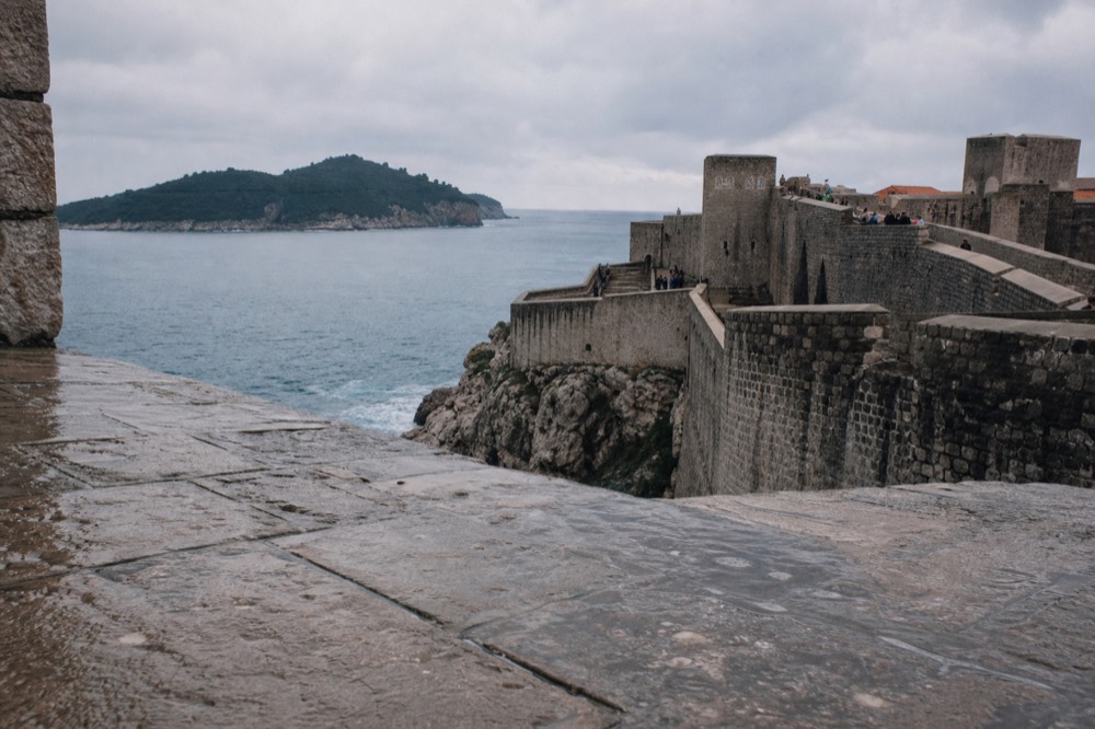 Stone fortress walls rising above the blue Adriatic Sea with a wooded island in the distance