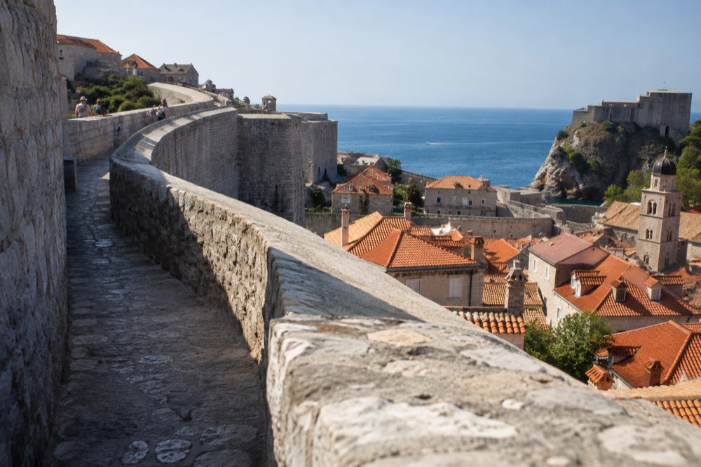 Stone city walls curving above red rooftops toward a blue sea horizon