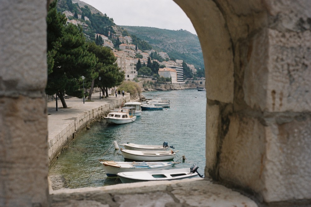 A stone quay with small boats moored in clear water beside a tree-lined promenade
