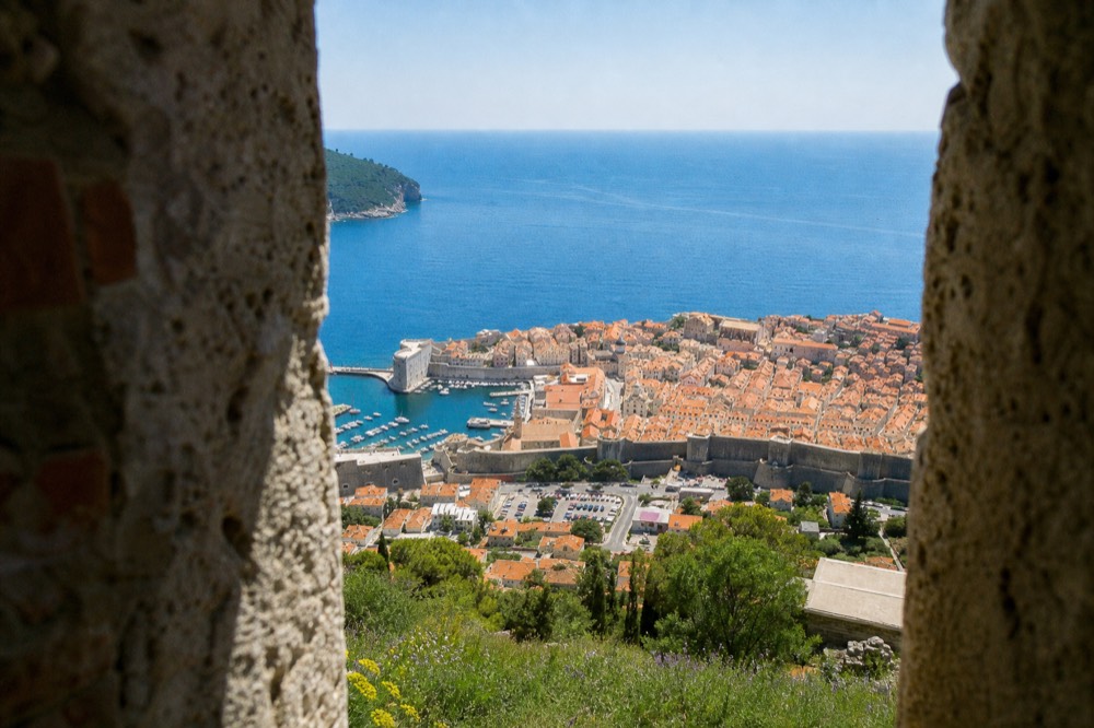 A walled medieval city with orange rooftops seen from a hillside above the sea