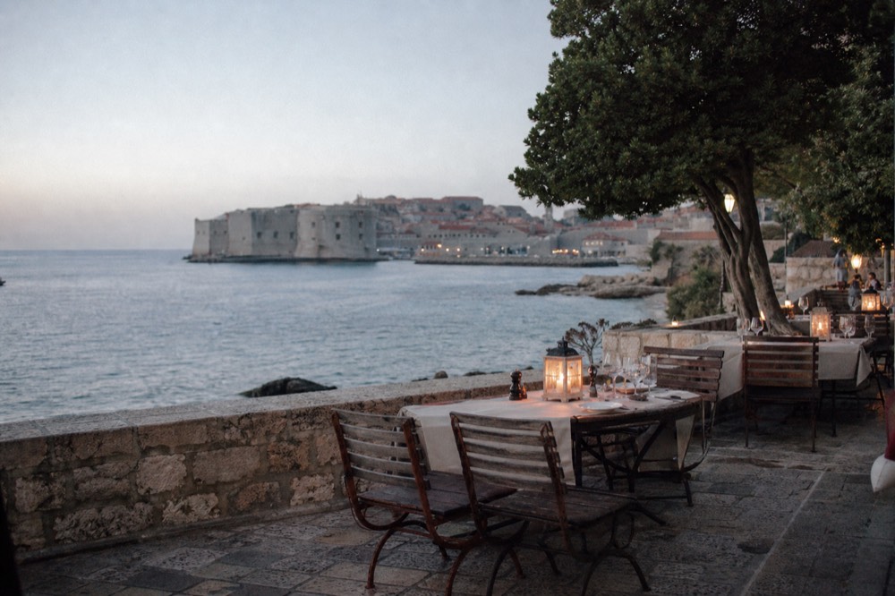 An outdoor dinner table beside the water at dusk with lanterns lit