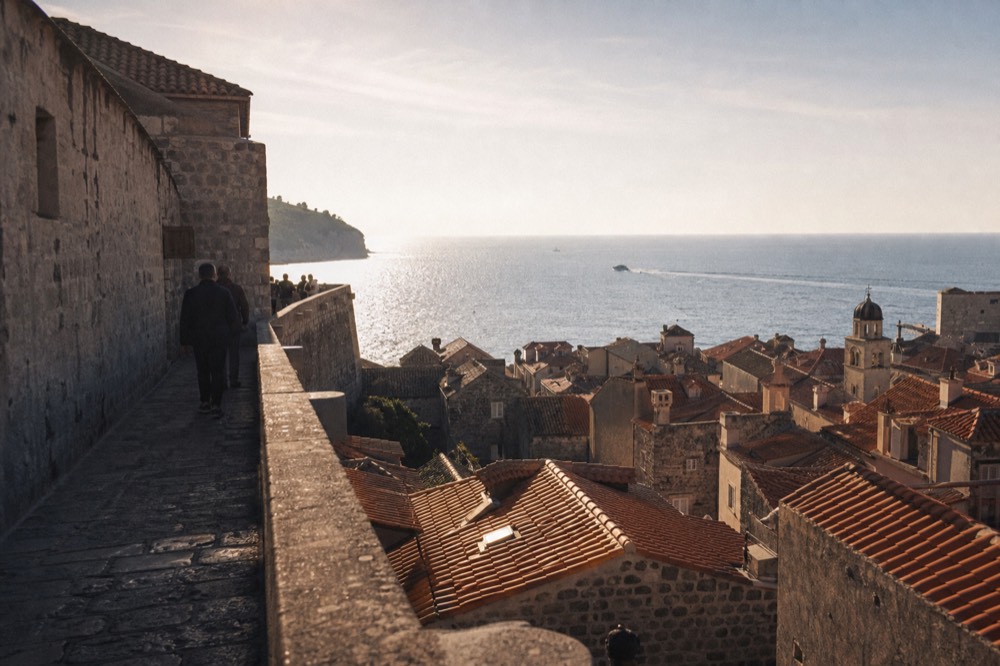 A stone city wall walkway above red rooftops with the sea on the horizon