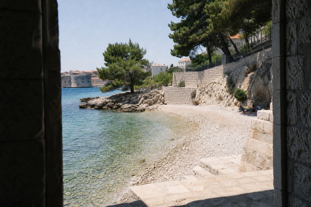 A pebble beach with clear shallow water and pine trees near a stone town