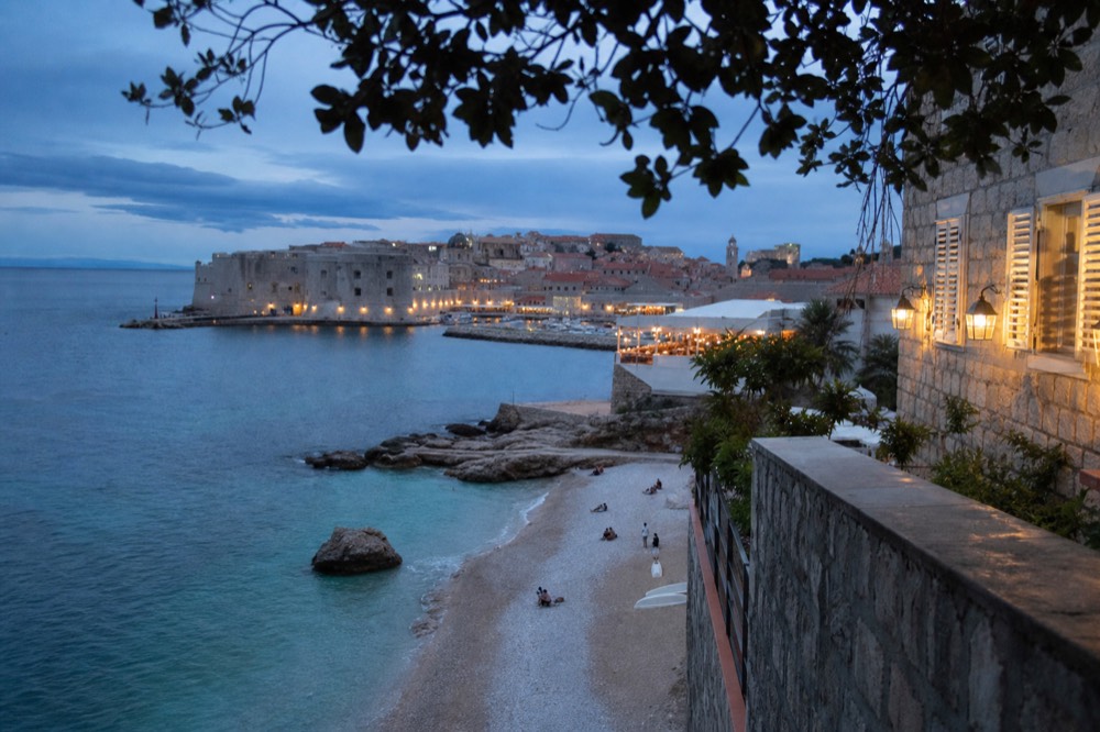 A pebble beach with clear turquoise water and a walled town beyond