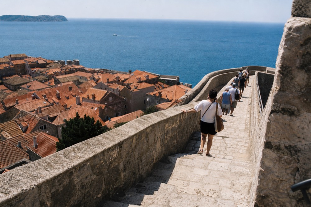 Walkers on a high stone city wall above red rooftops and the sea