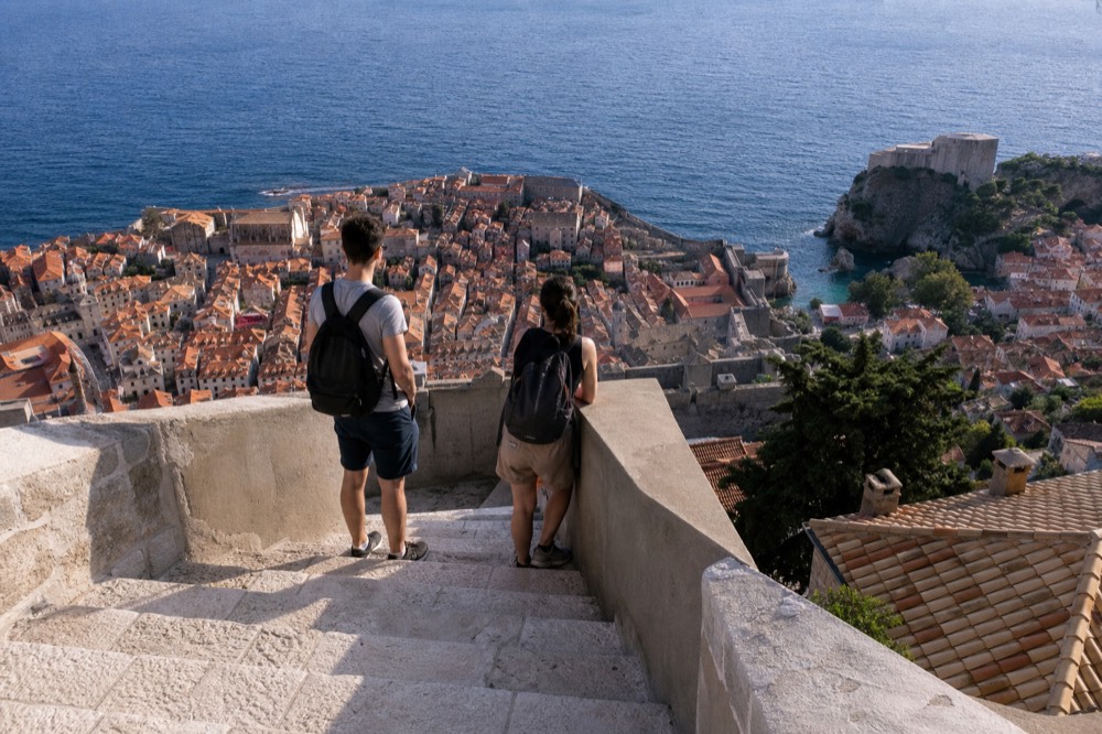 Two travellers with backpacks looking out over a coastal city from a high terrace