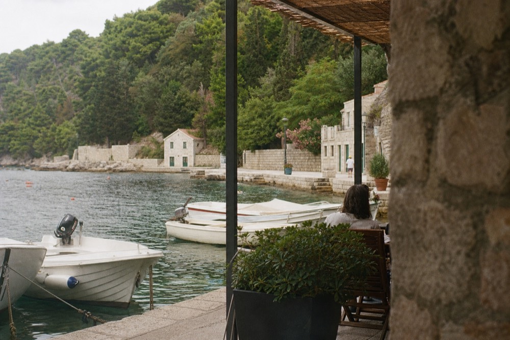 Shaded cafe terrace beside a stone quay with small boats moored in clear water