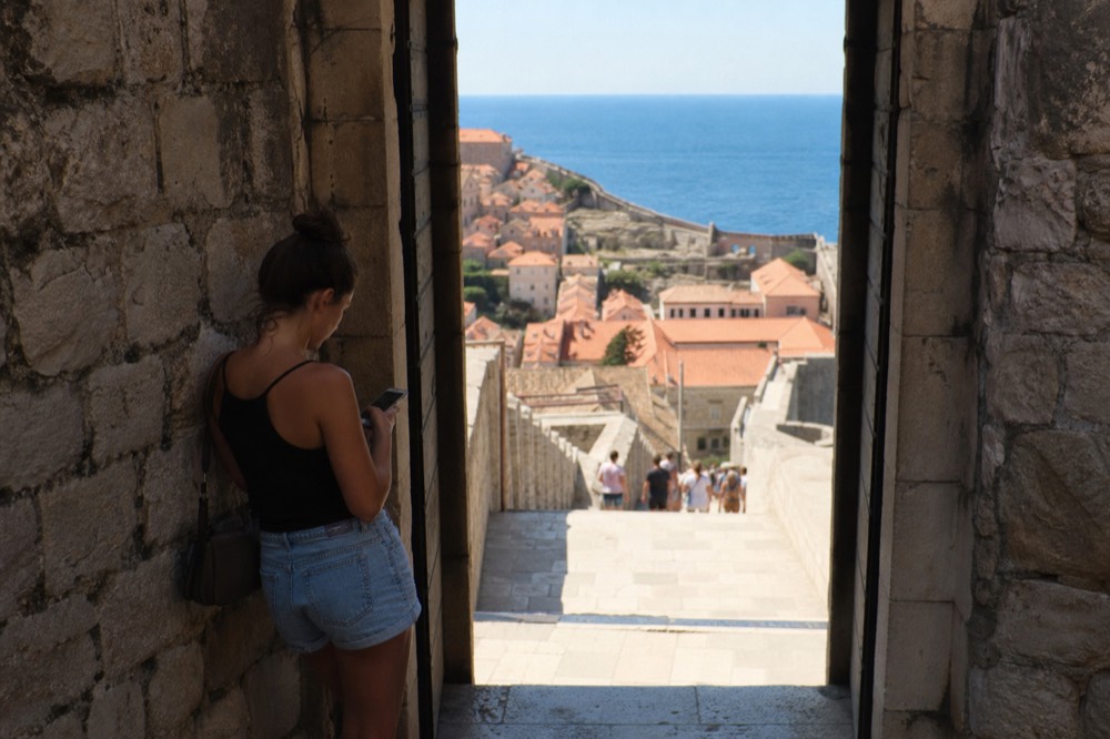 A traveller checking a phone at a stone gateway in Dubrovnik with a glimpse of the Adriatic beyond