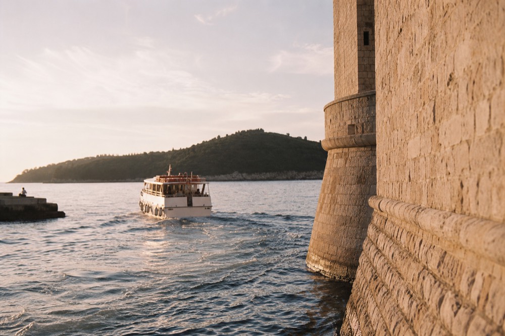 A small passenger boat departing Dubrovnik old harbour toward a green island