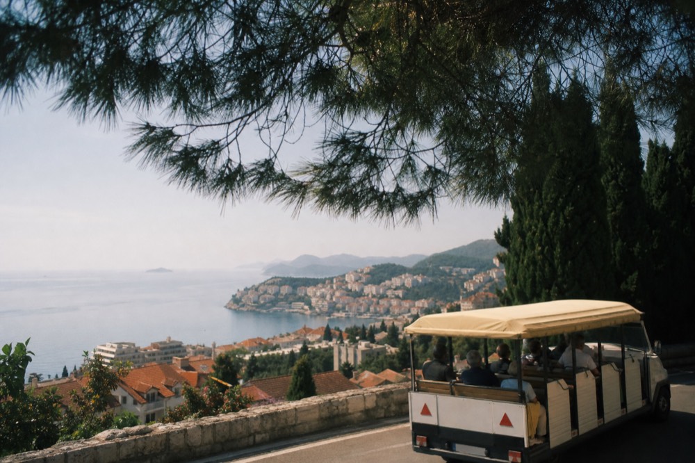 An open-sided sightseeing vehicle on a coastal road above a red-roofed town