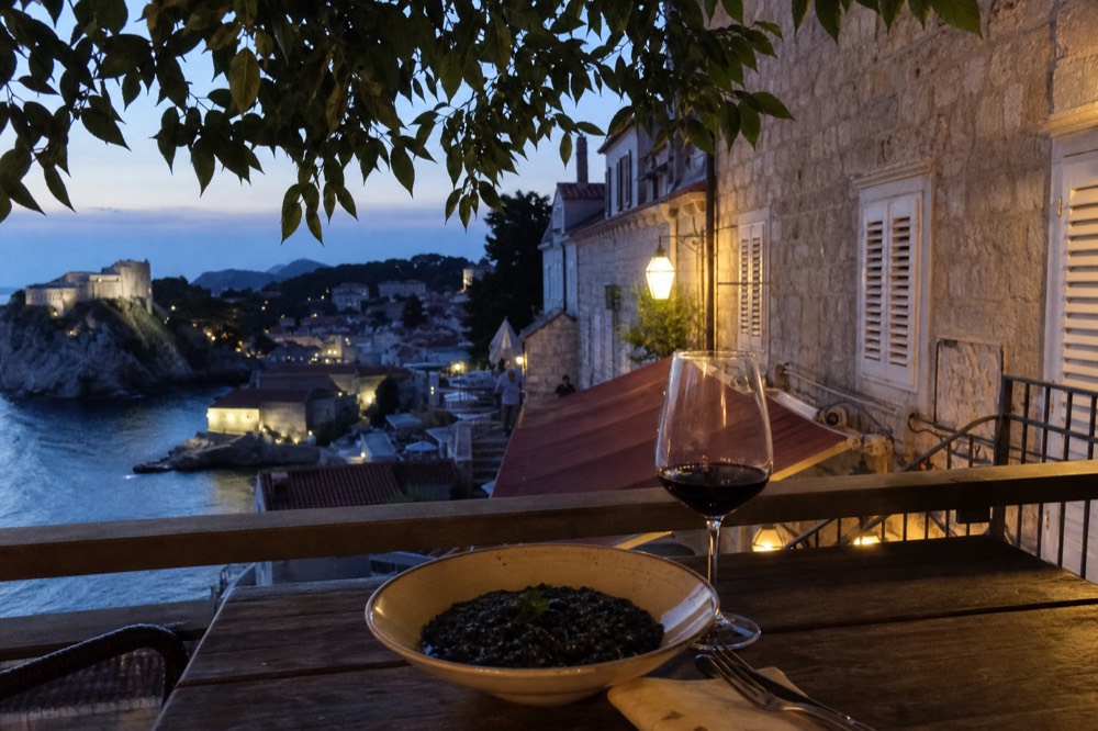 A bowl of black cuttlefish risotto on a rustic wooden table beside a glass of red wine