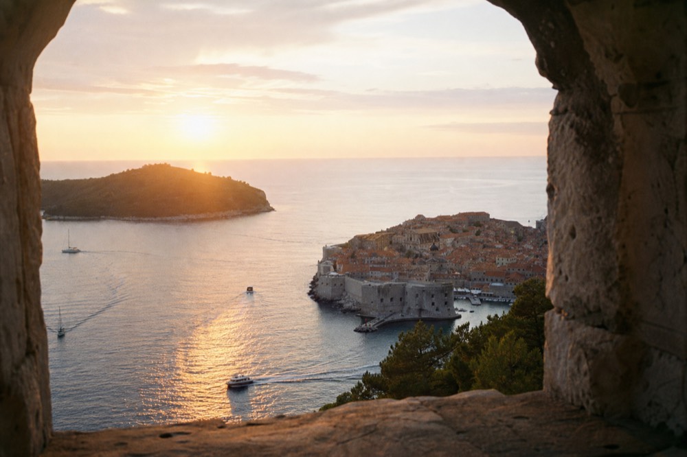 Panorama of a walled coastal city with red roofs and an island offshore at sunset