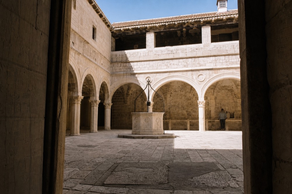 Sunlit arcade of a stone monastery courtyard with a well in the centre