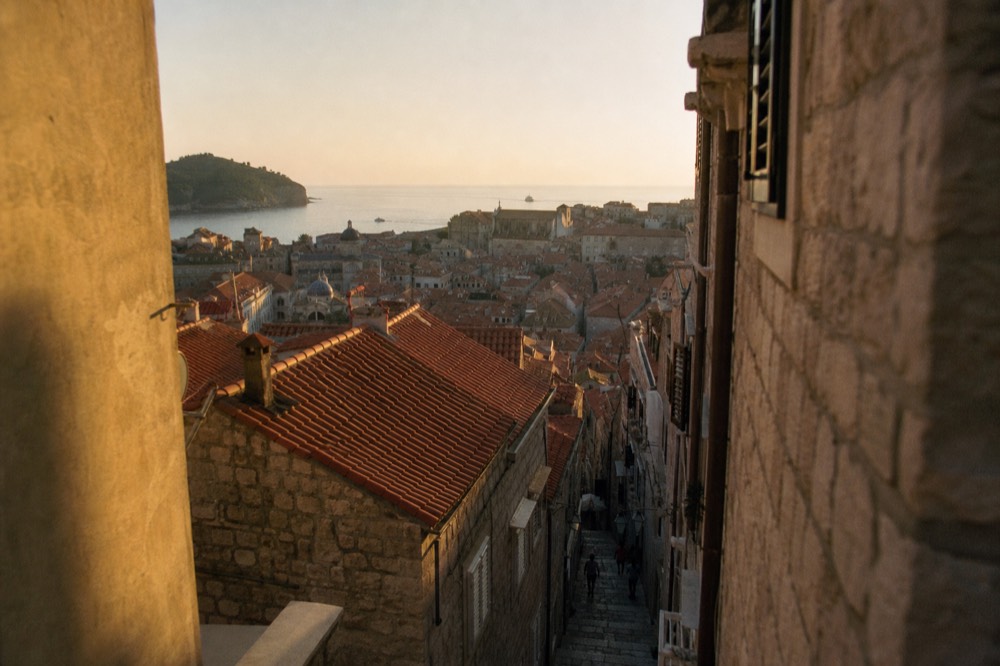 Walled coastal city at sunset with terracotta roofs and a darkening sky