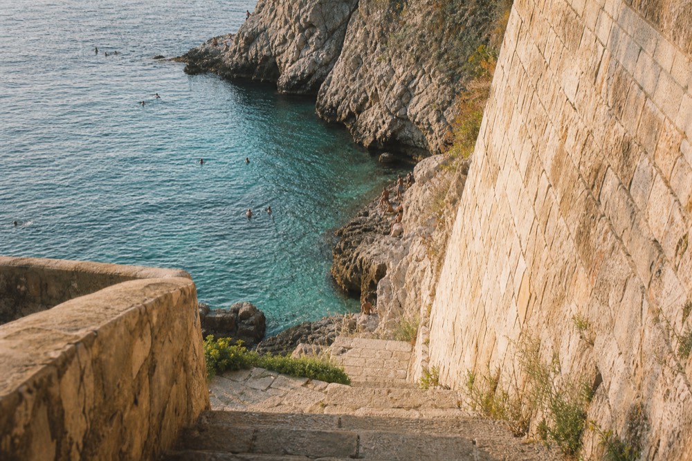 Swimmers in clear turquoise water below stone cliffs near Dubrovnik's city walls