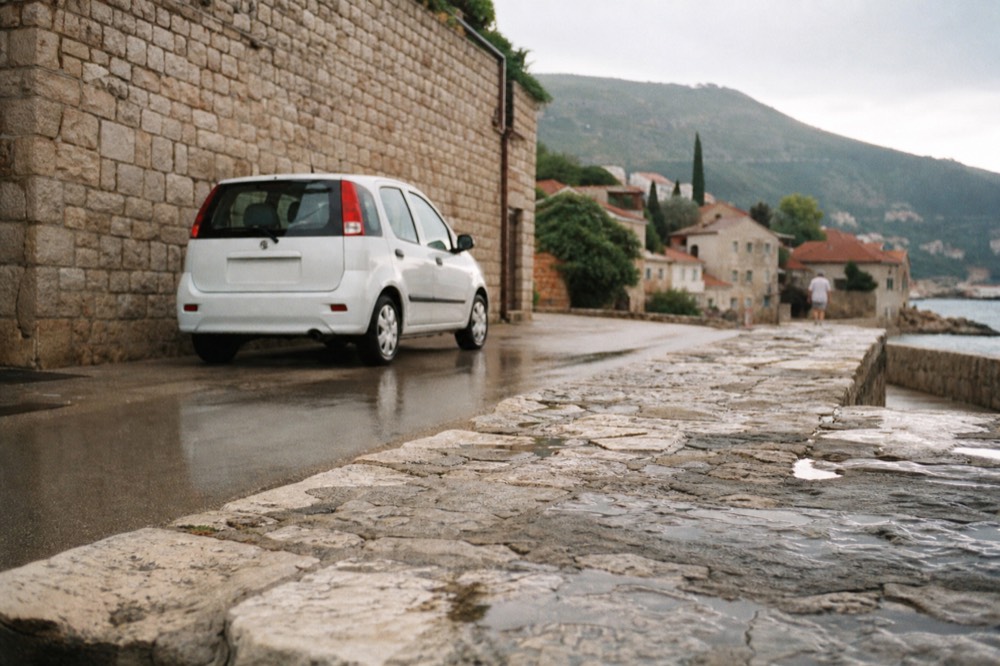A parked small rental car on a street beside stone buildings near the coast