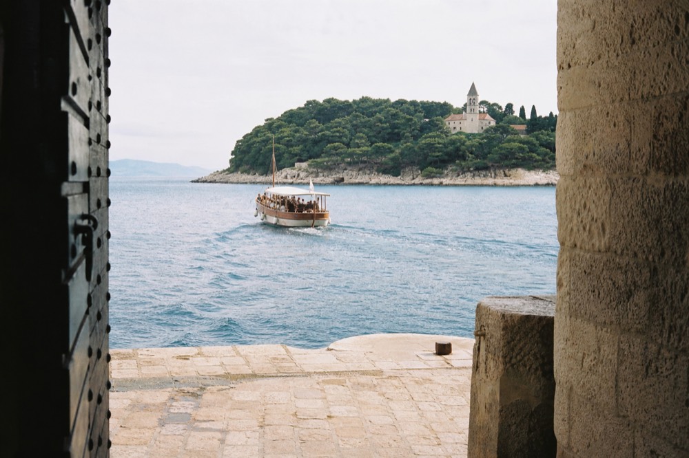 Wooden boat crossing calm blue water toward a green island with a monastery tower