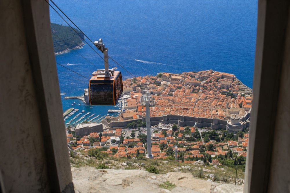 A cable car cabin ascending a steep hillside above a walled coastal city