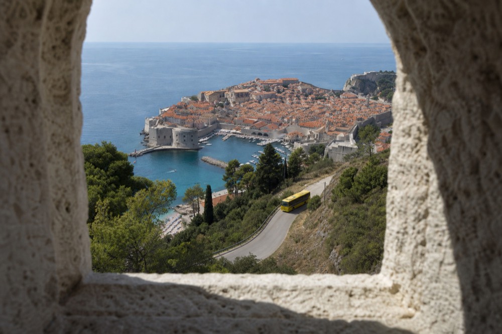 A yellow tour bus on a winding road above a terracotta rooftop old town by the sea