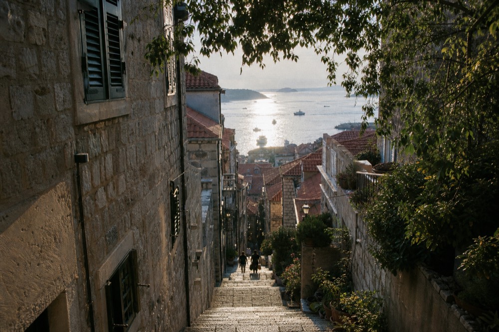 A narrow limestone street in an old town with stone staircases rising between shuttered buildings