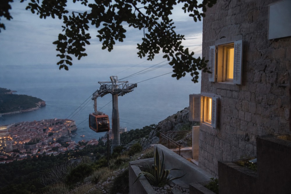 A cable car cabin ascending a steep hillside above a walled coastal city