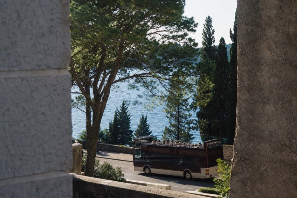 An open-top tour bus travelling a coastal road with pine trees and a blue sea beyond