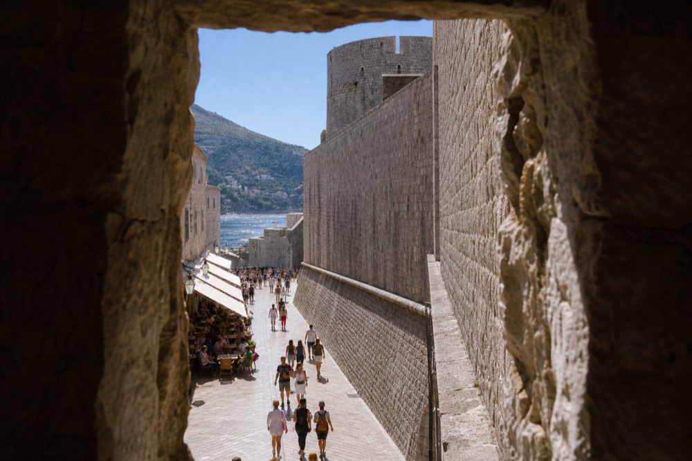 A stone fortress wall rising above a crowded marble street lined with cafe tables