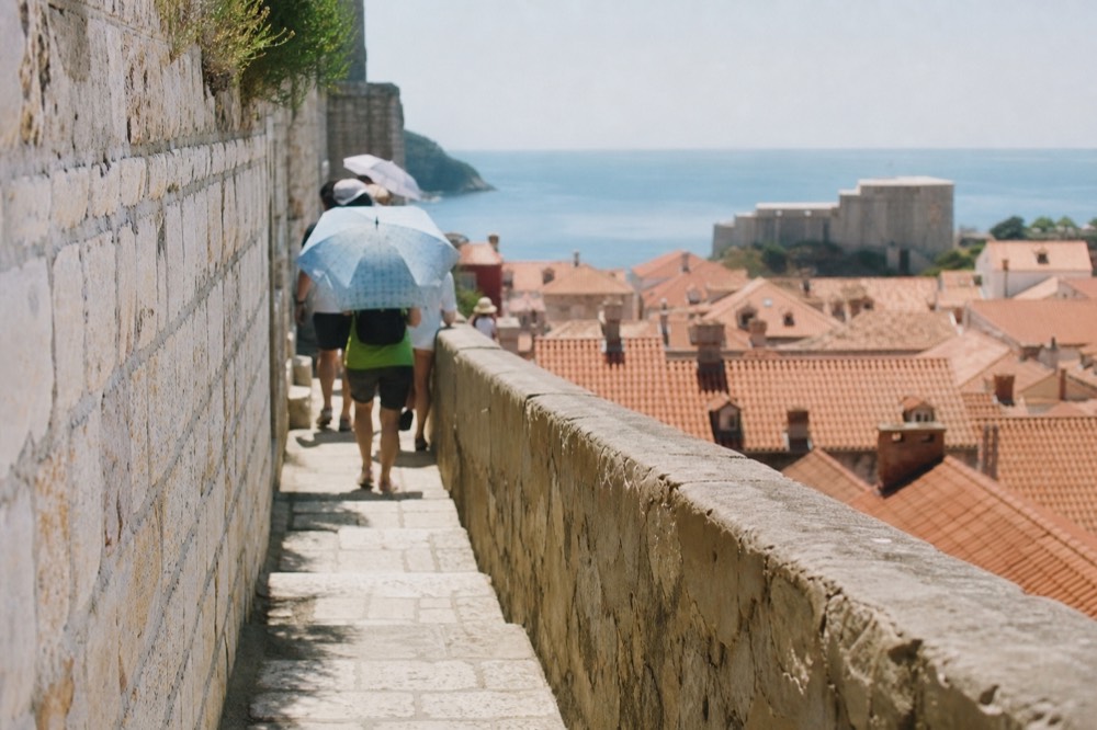 Tourists with umbrellas for shade walking along a high stone wall above a red-roofed city
