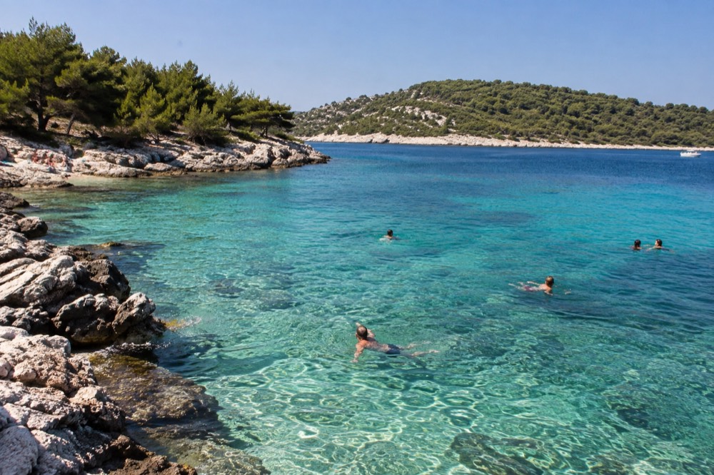 Swimmers in clear turquoise water beside a rocky island beach with green hills