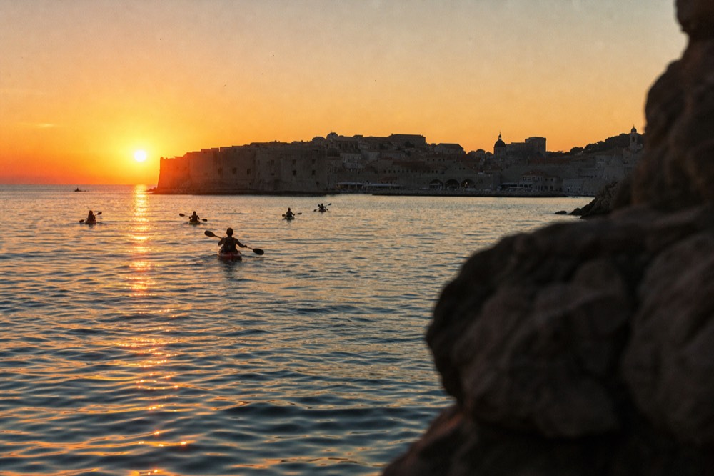 Kayaks on glassy water at sunset with Dubrovnik old town silhouetted against an orange sky