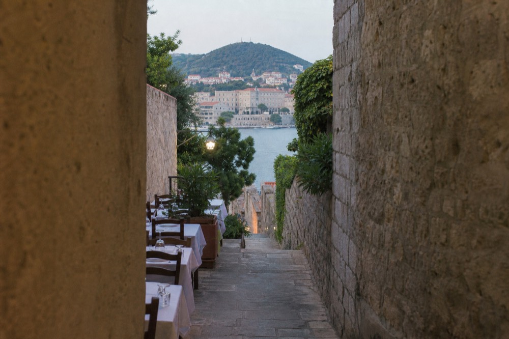 Wooden tables with white tablecloths set along a narrow stone alley at dusk