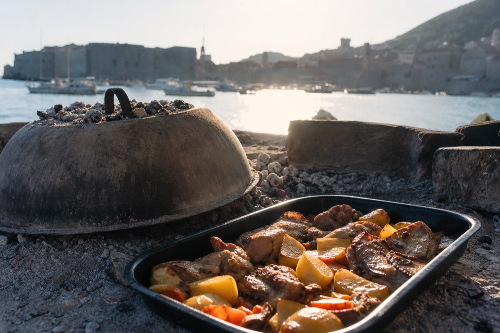 Domed baking bell over embers beside a tray of roasted vegetables and meat